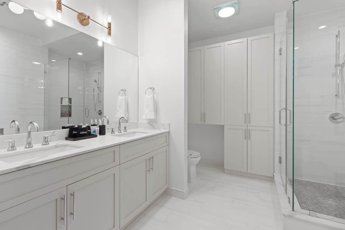 A modern bathroom with a large mirror, two sinks, a shower, and white cabinetry.