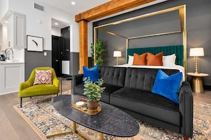 A modern hotel room with a black sofa, a green armchair, a round coffee table, and a bed with a green headboard.