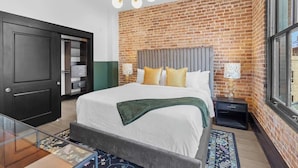 A bedroom with a brick wall, a bed with white bedding, a bedside table with a lamp, and a large window. - Game Room  Gym  Pool  Rooftop  Downtown Suite Hotel (New Orleans)