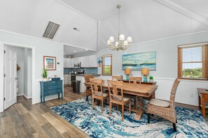 A spacious kitchen with white cabinets, a dining area with a wooden table and chairs, and a large window with blinds. - Second Wind: Oceanside with Views, Close to Beach and Lighthouse! (Corolla)