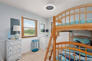 Uma cama beliche com estrutura de madeira e edredom azul e branco. Há uma cômoda branca com gavetas, uma pequena mesa de cabeceira azul e uma luminária sobre a cômoda. É visível uma janela com persianas, e há um luminário no teto.