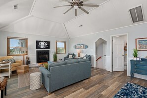A spacious living room with a fireplace, a ceiling fan, and a television mounted on the wall. - Second Wind: Oceanside with Views, Close to Beach and Lighthouse! (Corolla)