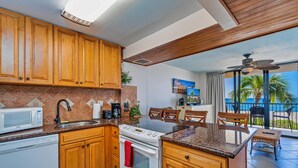 Una cocina con armarios de madera, microondas y estufa. Hay un comedor con sillas y vistas al océano.