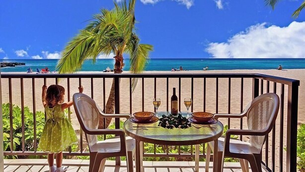 Un balcón con una mesa lista para dos personas, con vistas a una playa con palmeras y un cielo azul despejado.