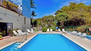 Una piscina rectangular con agua azul cristalina, rodeada de sillas de descanso blancas y un edificio con balcón.