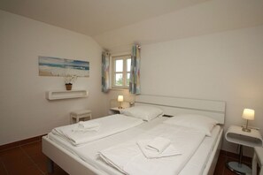 A hotel room with a double bed, two bedside tables with lamps, a small window with curtains, a shelf with a plant, and a painting on the wall. - Vacation apartment close to the beach with private terrace - Feriendorf Bakenberg FeWo C27 - (Dranske)