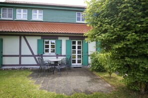 A two-story building with a tiled roof, green shutters, and a small patio with a table and chairs.