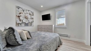 Un dormitorio con cama, televisión, ventana con persianas y una obra de arte colgada en la pared.