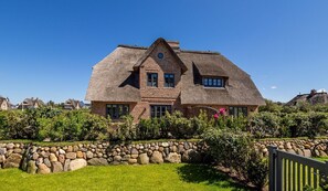 A large, thatched-roof house with a stone wall in front, surrounded by greenery.