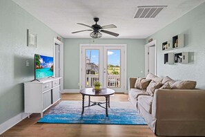 A living room with a television, a ceiling fan, a dining table, a sofa, and a view through a glass door.