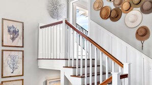 Un escalier avec des rampes blanches et des mains courantes en bois, entouré d’œuvres d’art encadrées et d’une collection de chapeaux accrochés au mur.