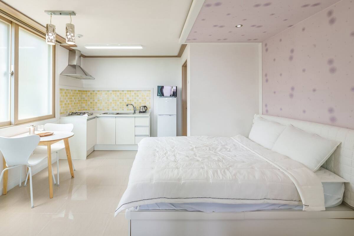 A modern hotel room with a bed, a small dining table, and chairs. There is a kitchenette with a stove, sink, and refrigerator.