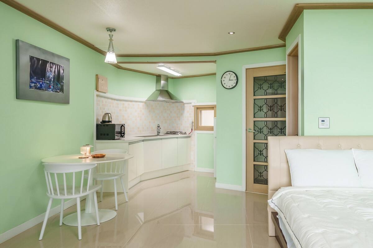 A room with a bed, a small kitchenette, a dining area with a table and chairs, and a clock on the wall.