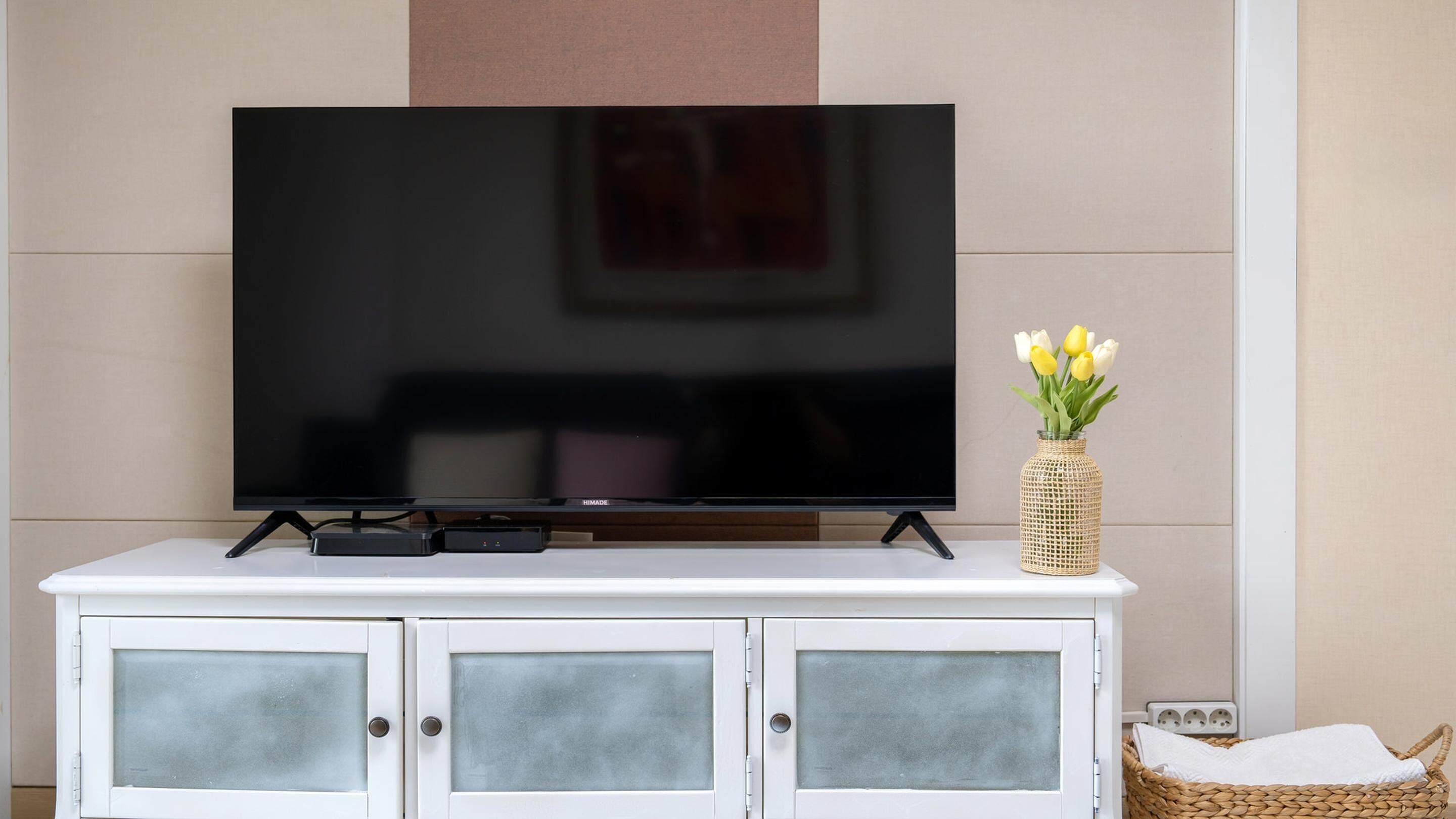 A flat-screen TV on a white cabinet with frosted glass doors, a woven vase with yellow flowers, and a woven basket with a white towel.