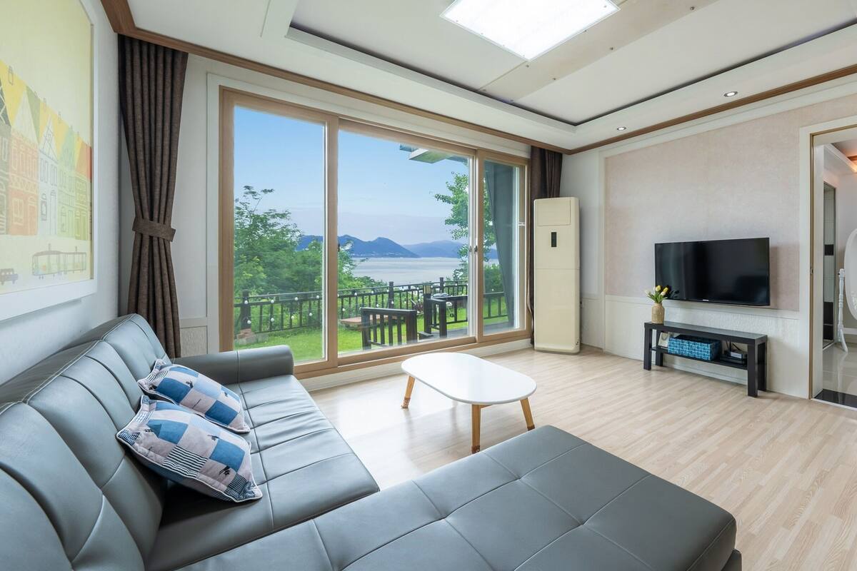A modern living room with a sofa, a coffee table, and a TV. There is a large window offering a view of a balcony and mountains.