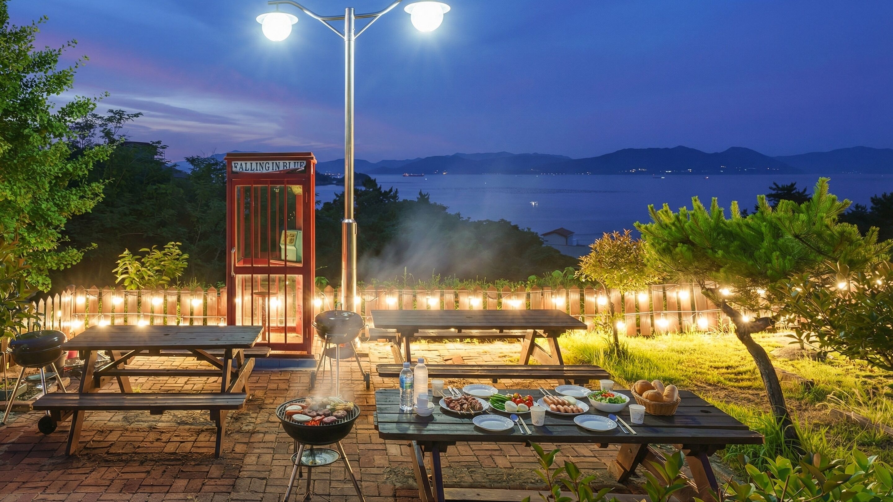 An outdoor dining area with a grill, tables, and a view of the sea at dusk.
