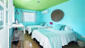 Ein Zimmer mit zwei Betten, einem fensterbelaubten Fenster, einer Wand-Klimaanlage, einem gewebten Korb an der Wand und einem kleinen Tisch mit einer Lampe.