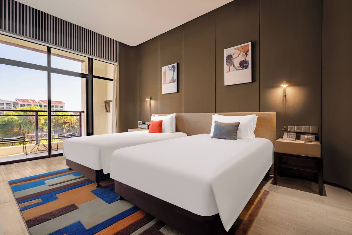 A modern hotel room with two beds, a large window with a view, and a small bedside table with a lamp.