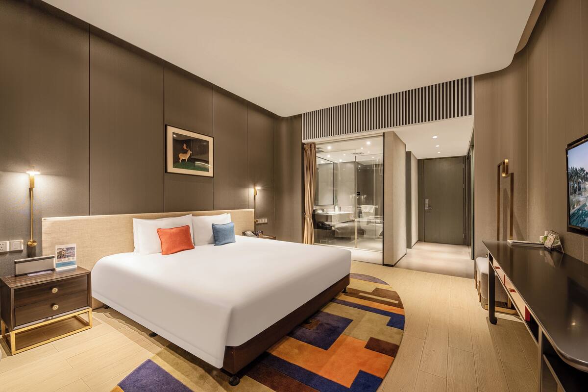A modern hotel room with a large bed, bedside tables, a desk, and a glass-enclosed bathroom.