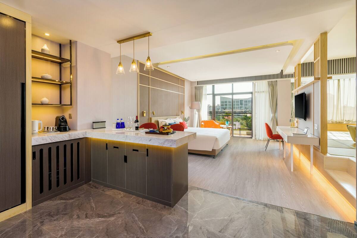A modern hotel room with a kitchen area, a bed, a desk, and a large window offering a city view.
