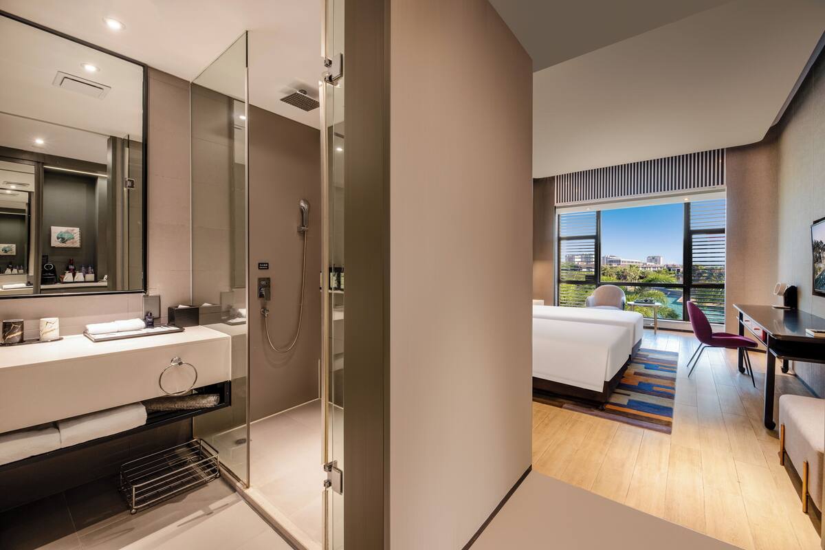 A modern hotel room with a glass-enclosed shower, a large bed, a desk, and a view of the city.