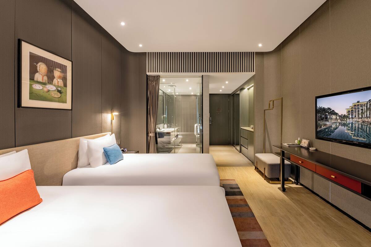 A modern hotel room with a large bed, a desk, a television, and a view of the city through the window.