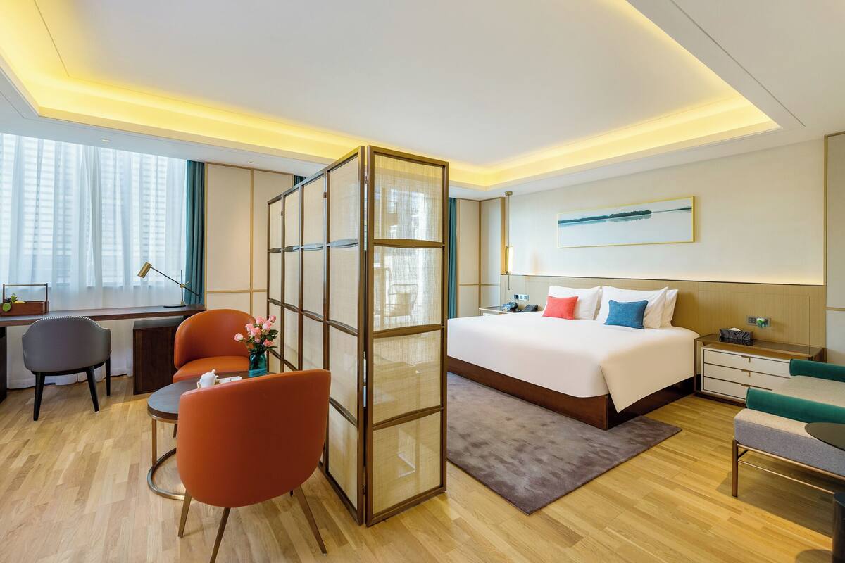 A modern hotel room with a large bed, a desk with a chair, a glass partition, and a view of the outdoors.