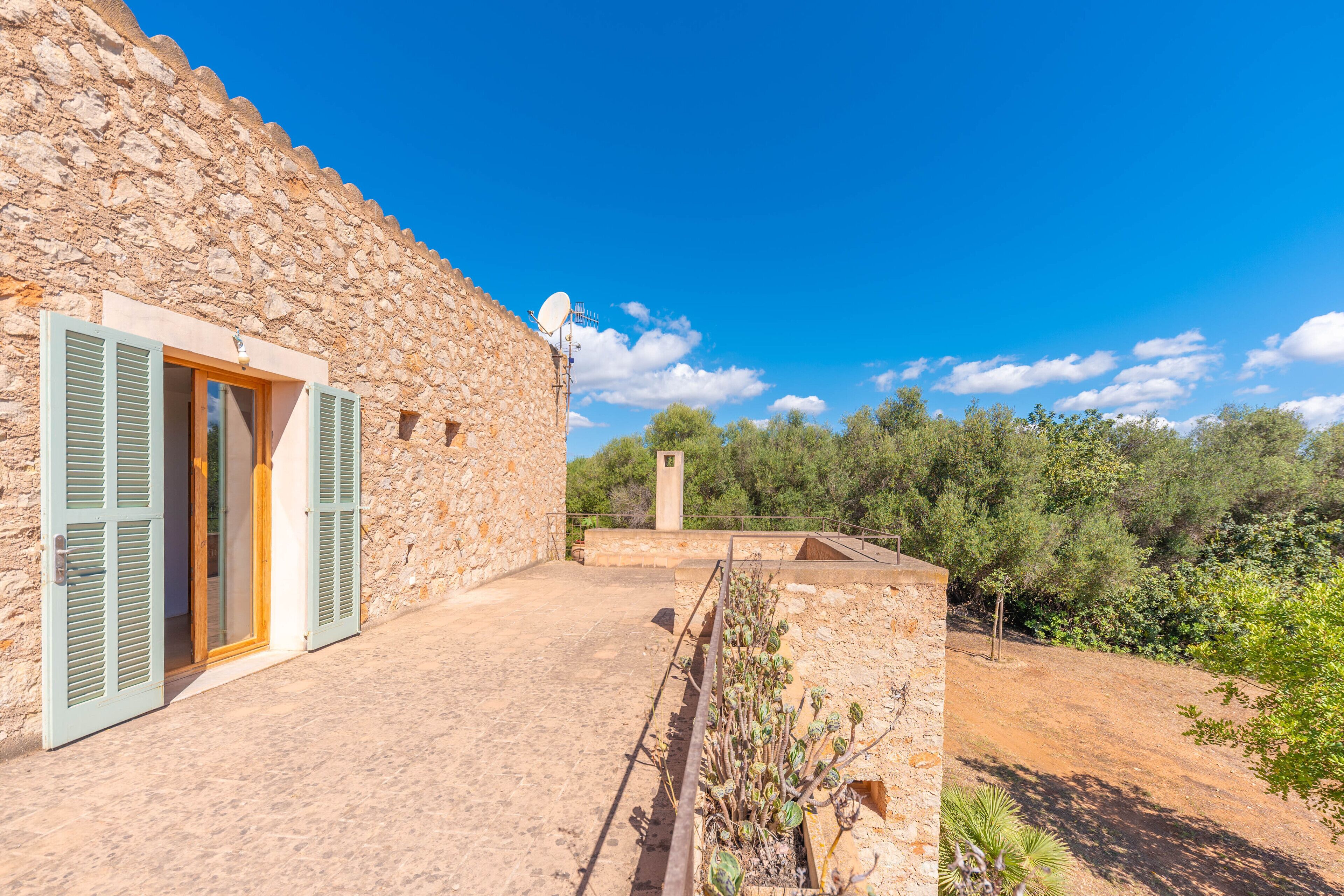 Un edificio de piedra con puerta y contraventanas, una antena parabólica y vistas a árboles y cielo.