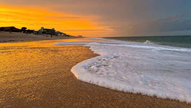 Uma praia com pôr do sol, ondas e edifícios ao fundo.