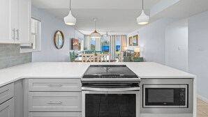 A modern kitchen with white cabinets, a built-in stove, oven, and microwave. There are pendant lights, a dining area with a table and chairs, and a view of the outdoors. - 2 BR Beach Condo, Boutique Resort, 1st Fl Walkout, Steps to Pool & Beach Sleep 6 (Satellite Beach)