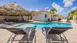 A pool area with lounge chairs, a thatched-roof structure, and a small building. - Colonial Modern Villa Sleep 14-Pool, Palapa, Court (Noord)