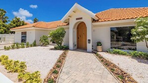 A building with a tiled roof, a central entrance with an arched doorway, and a gravel driveway with plants. - Colonial Modern Villa Sleep 14-Pool, Palapa, Court (Noord)