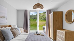 A bedroom with a bed, a bedside table, a dresser, and a large window with a view of greenery. - 6 The Vista - Hot tub/ Pet friendly/ On-site pool/ EV Charger/ Ecohome (Woolacombe)