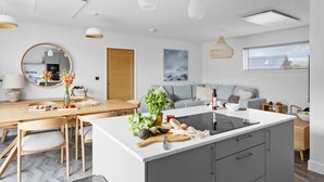 A modern kitchen with a central island, wooden dining table, and a grey sofa. - 6 The Vista - Hot tub/ Pet friendly/ On-site pool/ EV Charger/ Ecohome (Woolacombe)