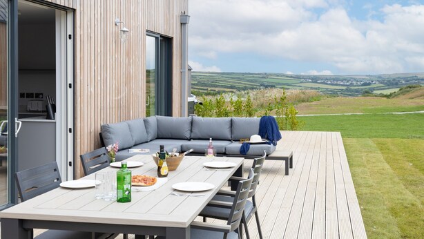 An outdoor dining area with a wooden deck, a grey sofa, and a table set for two. - 6 The Vista - Hot tub/ Pet friendly/ On-site pool/ EV Charger/ Ecohome (Woolacombe)