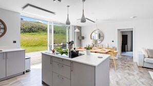 A modern kitchen with a large island, a dining area with a wooden table, and a view of the outdoors. - 6 The Vista - Hot tub/ Pet friendly/ On-site pool/ EV Charger/ Ecohome (Woolacombe)
