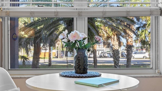 A round table with a vase of flowers and a book, a window with a view of palm trees and a street. - Updated! Beautiful Decor! Walk to Beach & Eats (St. Augustine)