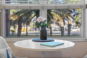 A round table with a vase of flowers and a book, a window with a view of palm trees and a street. - Updated1 Beautiful Decor! Walk to Beach & Eats (St. Augustine)