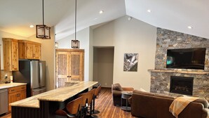 Une cuisine moderne avec des armoires en bois, un réfrigérateur en acier inoxydable, un coin bar avec des tabourets, une cheminée en pierre et un grand téléviseur fixé au mur.