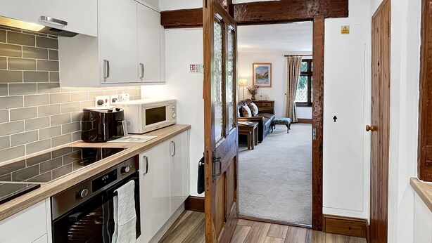 A narrow kitchen with white cabinets, a microwave, and a stove. A wooden door leads to a living area with a sofa and a table. - Gardener Cottage near Martinhoe (Parracombe)