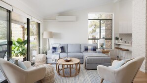 A modern living room with a grey sofa, two armchairs, a round wooden coffee table, and a striped rug. There is a large window with a view of a brick wall and greenery outside.