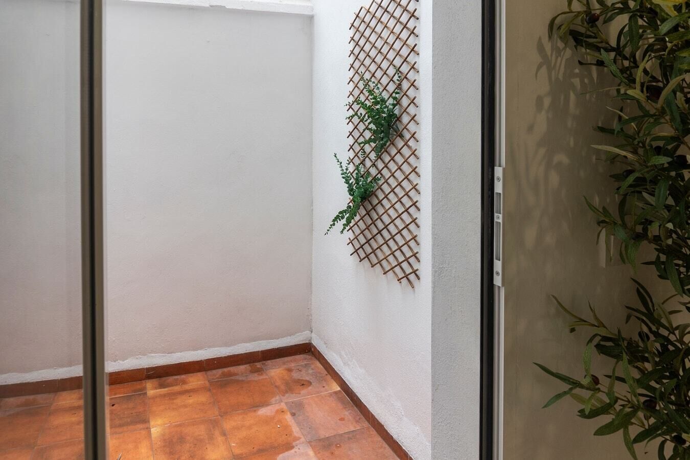 Un petit espace clos doté d’un sol carrelé, d’un mur blanc et d’une structure en treillis de bois ornée de plantes vertes.