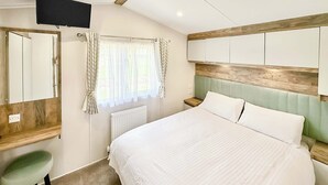 Un dormitorio con cama, televisión montada en la pared, ventana con cortinas, radiador y cabecero de madera.