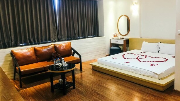 A hotel room with a sofa, a small table, a bed with a heart-shaped decoration, a mirror, and a window with curtains. - Hoang Gia Hotel 150 Nghi Tam HN-BayLux (Hanoi)