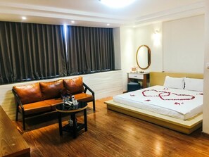 A hotel room with a sofa, a small table, a bed with a heart-shaped decoration, a mirror, and a window with curtains. - Hoang Gia Hotel 150 Nghi Tam HN-BayLux (Hanoi)