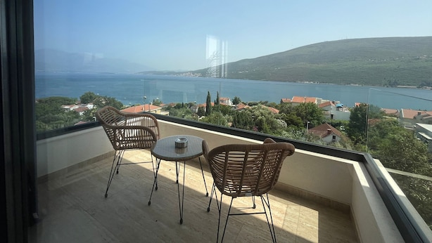 A balcony with two wicker chairs and a small table, overlooking a body of water and a bridge.