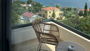 A balcony with a wicker chair and a small table, overlooking a coastal town with buildings and a clear view of the sea.