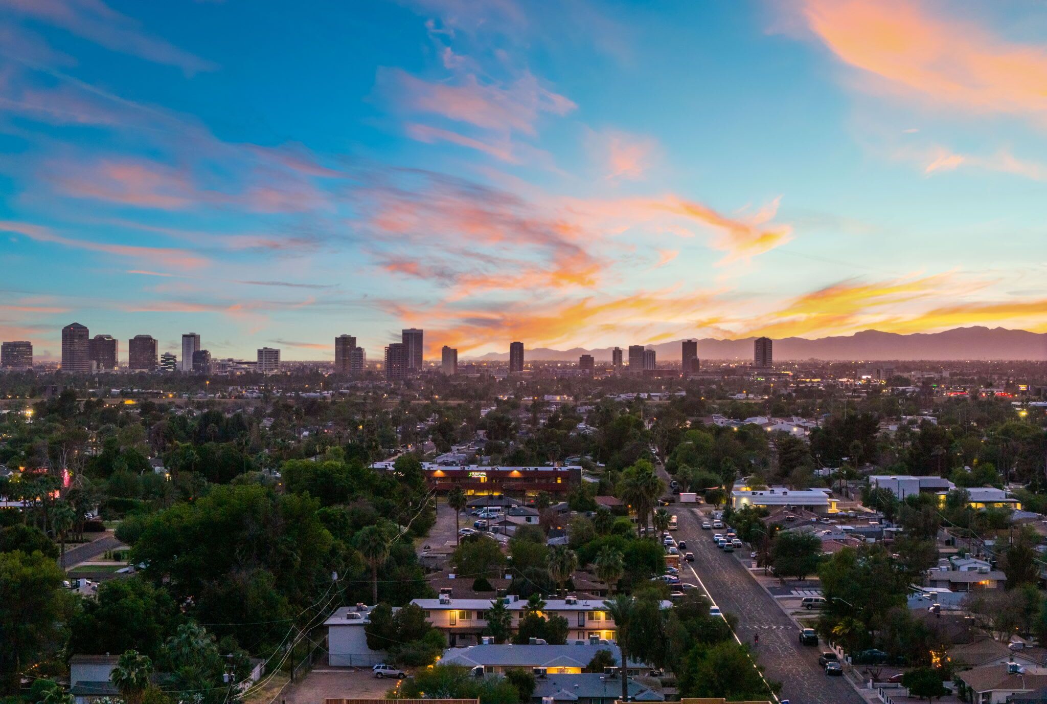 A cityscape at sunset with a skyline of buildings, a road with vehicles, and surrounding greenery.