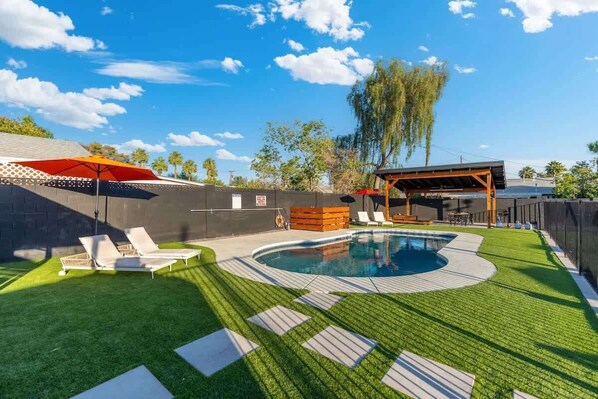 A backyard with a swimming pool, artificial grass, lounge chairs, and a covered seating area.