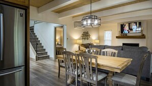 Une cuisine moderne avec un réfrigérateur en acier inoxydable, une table à manger en bois et des chaises. On y trouve un escalier menant à un autre niveau, un canapé et un téléviseur fixé au mur.